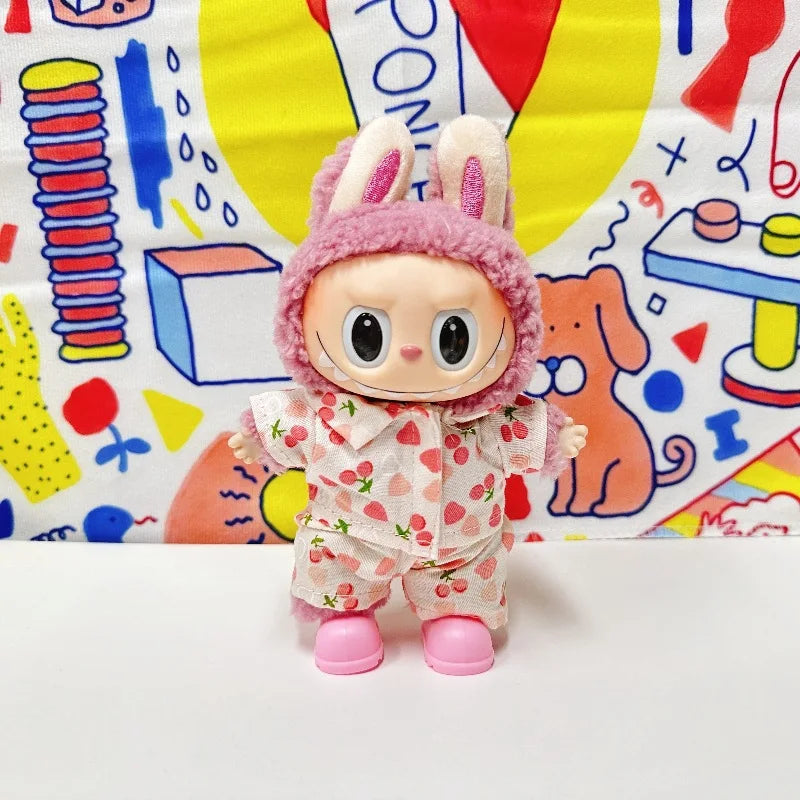 A Labubu figure wearing a light-colored two-piece pajama set with a red strawberry print. The figure also has a fluffy pink hood with bunny ears on its head and is wearing pink shoes.