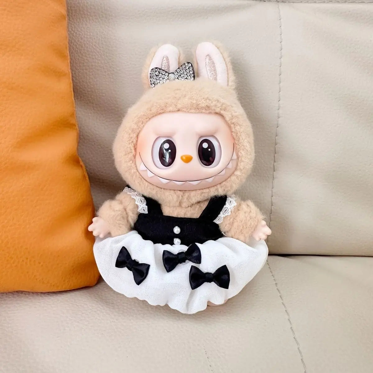 A Labubu figure wearing an elegant black dress with a white skirt and three black bows. The figure is sitting on a white leather sofa next to an orange cushion. A matching black bow with rhinestone accents is on the figure's head.

